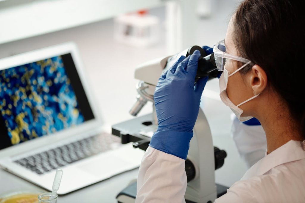 Scientist in lab coat examines samples using a microscope beside laptop, highlighting modern research techniques.